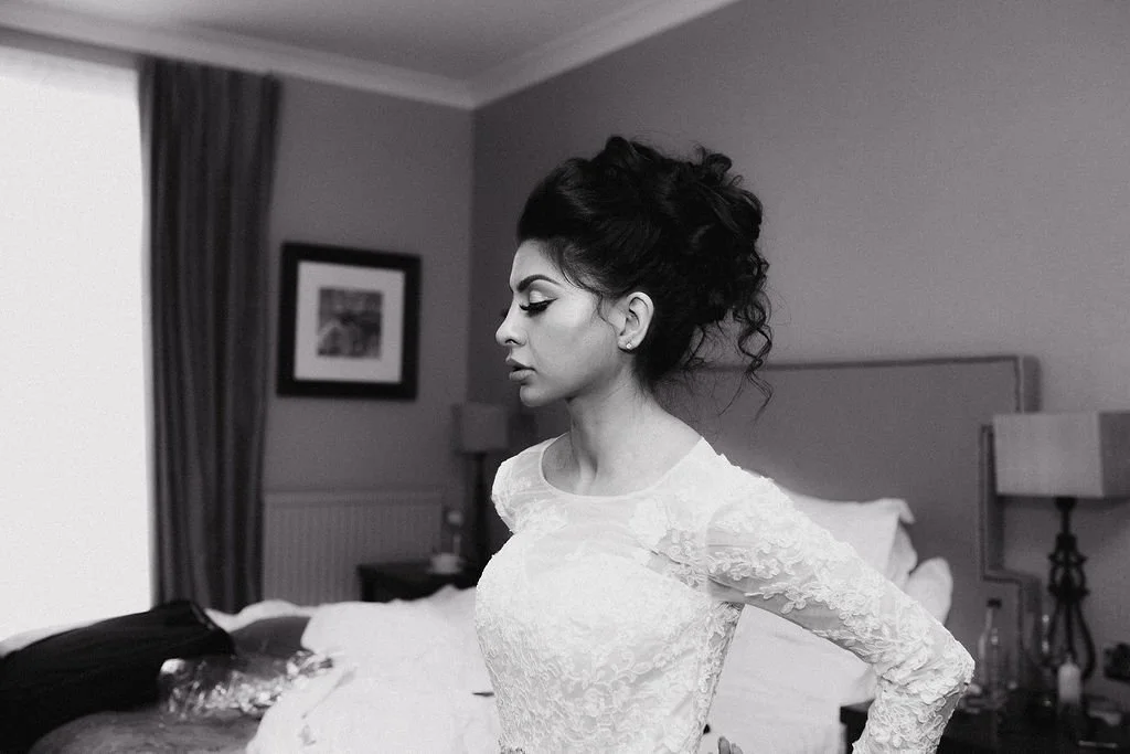 A woman with dark, curly hair in an updo, wearing a lace top, standing in a hotel room.