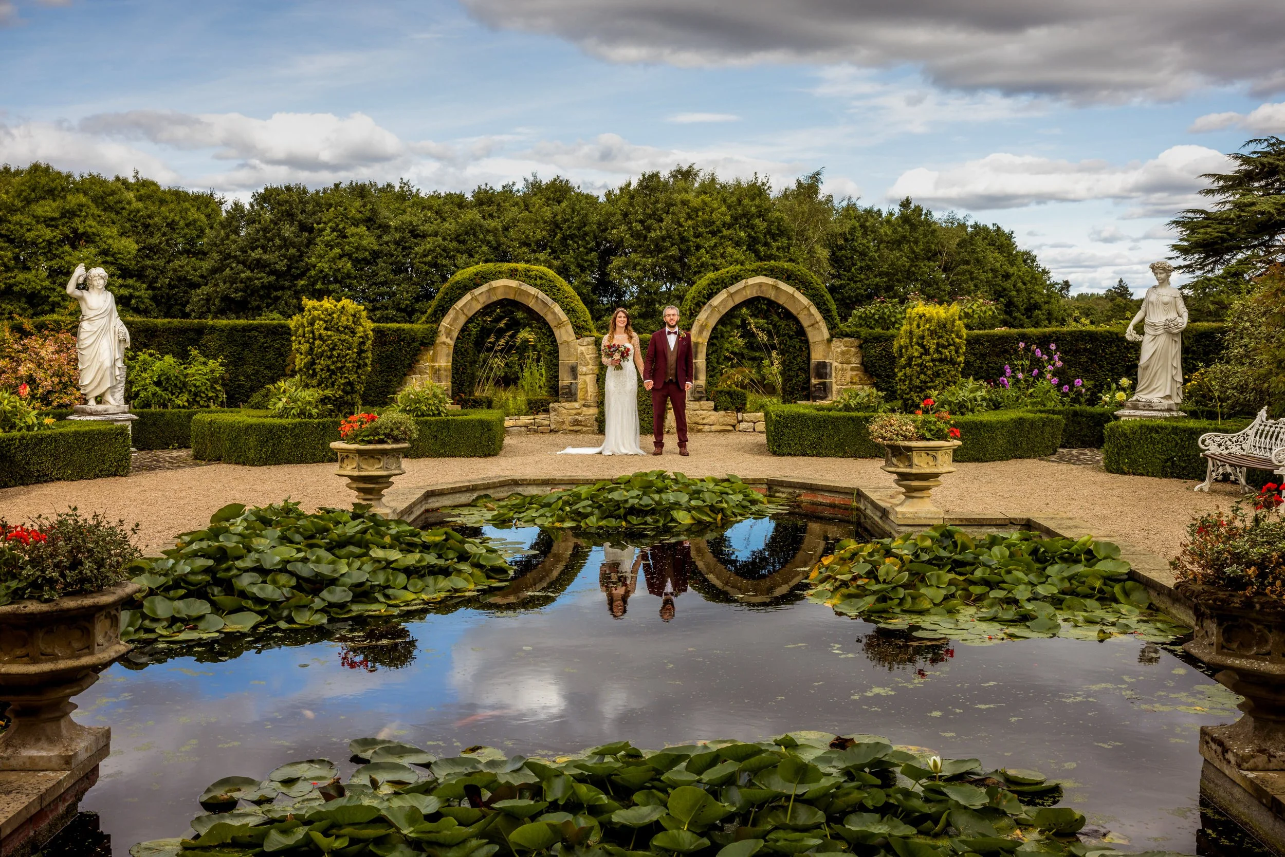 A bride and groom holding hands in a garden near a pond with lily pads, surrounded by statues, lush greenery, and stone arches.