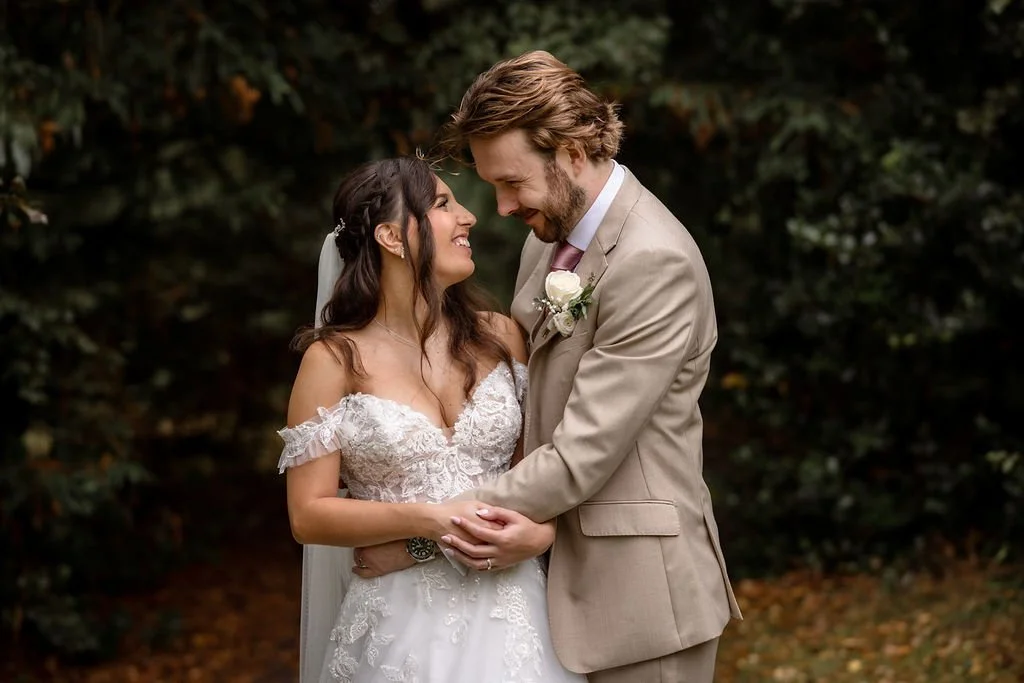 A smiling bride and groom holding hands and looking into each other's eyes outdoors, with trees and foliage in the background.