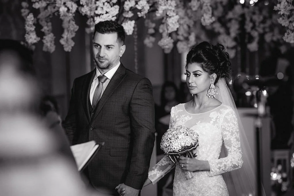 Black and white photo of a bride and groom during their wedding ceremony, with the bride holding a bouquet of flowers and an officiant reading from a book.
