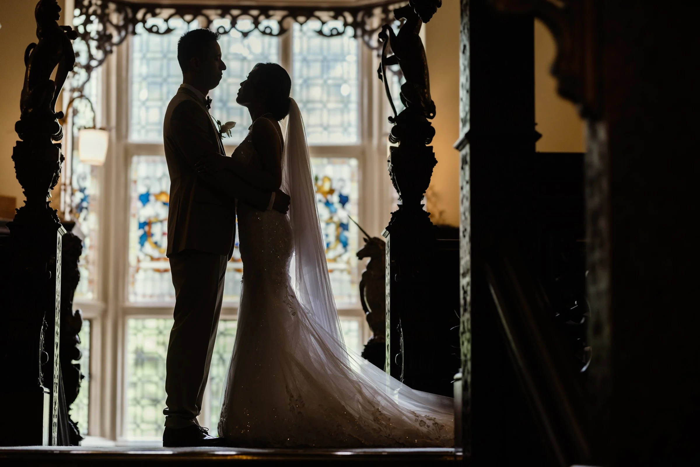 Silhouette of a bride and groom facing each other in a church interior with large window behind them, ornate curtains, and wooden sculptures on either side.