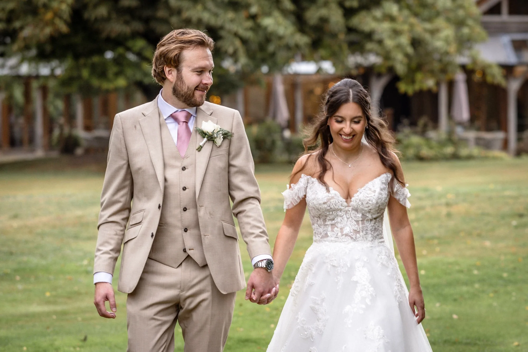 A bride and groom walking hand in hand on a grassy lawn, smiling and looking at each other, outdoors with trees and buildings in the background.