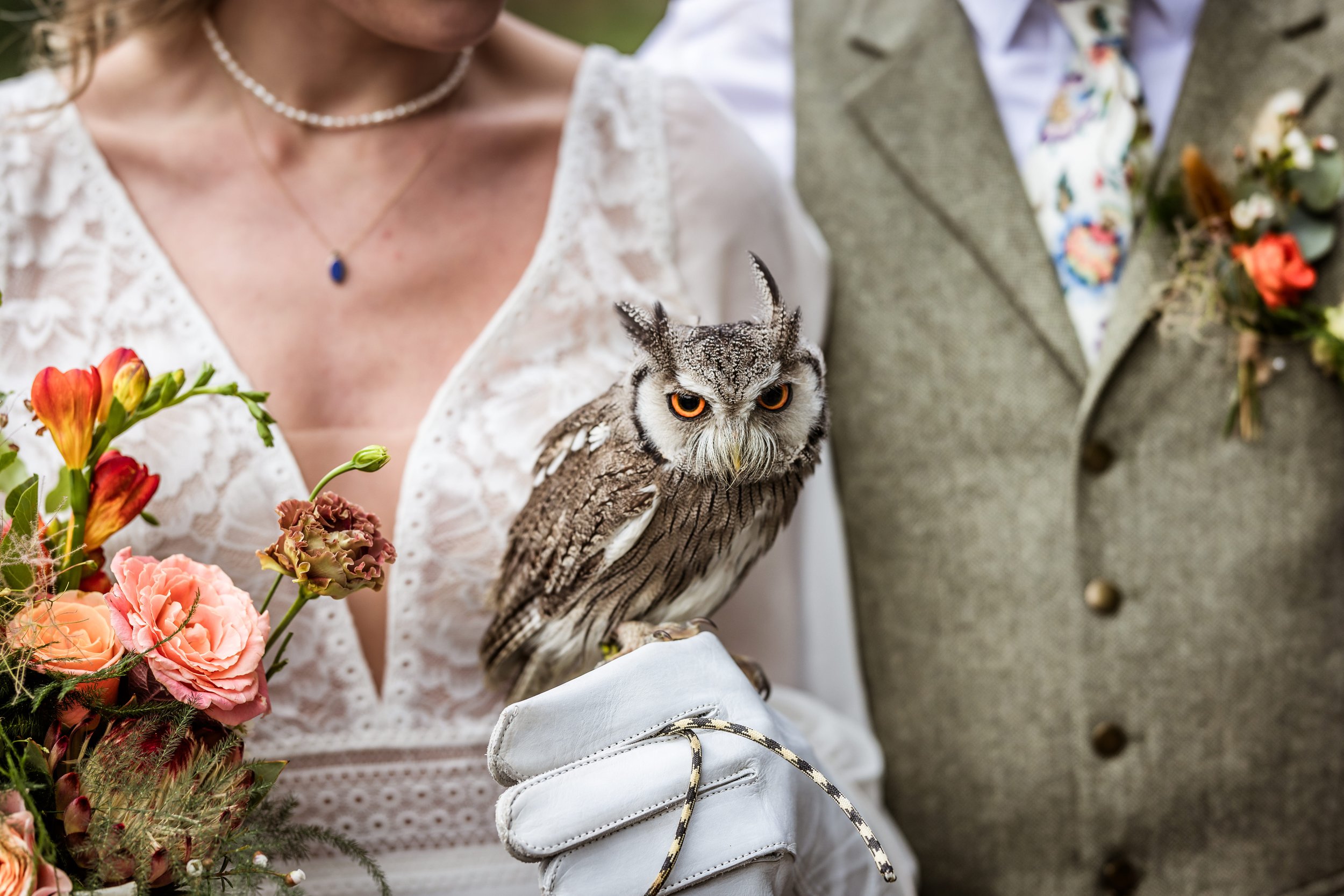 A woman holding a grey owl with orange eyes at a wedding, with a man in a grey suit and floral tie standing beside her. The woman is wearing a white lace dress, a pearl necklace, and gloves, and is holding a bouquet of pink, peach, and red flowers.