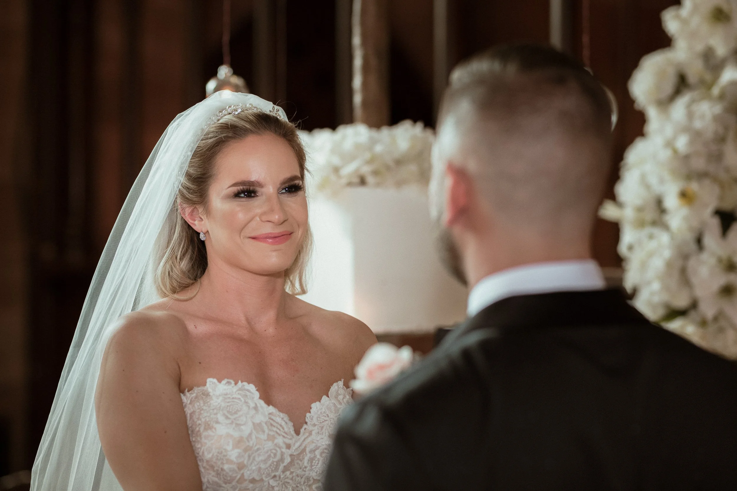 A bride and groom during their wedding ceremony, with the bride smiling at the groom in a close-up shot.