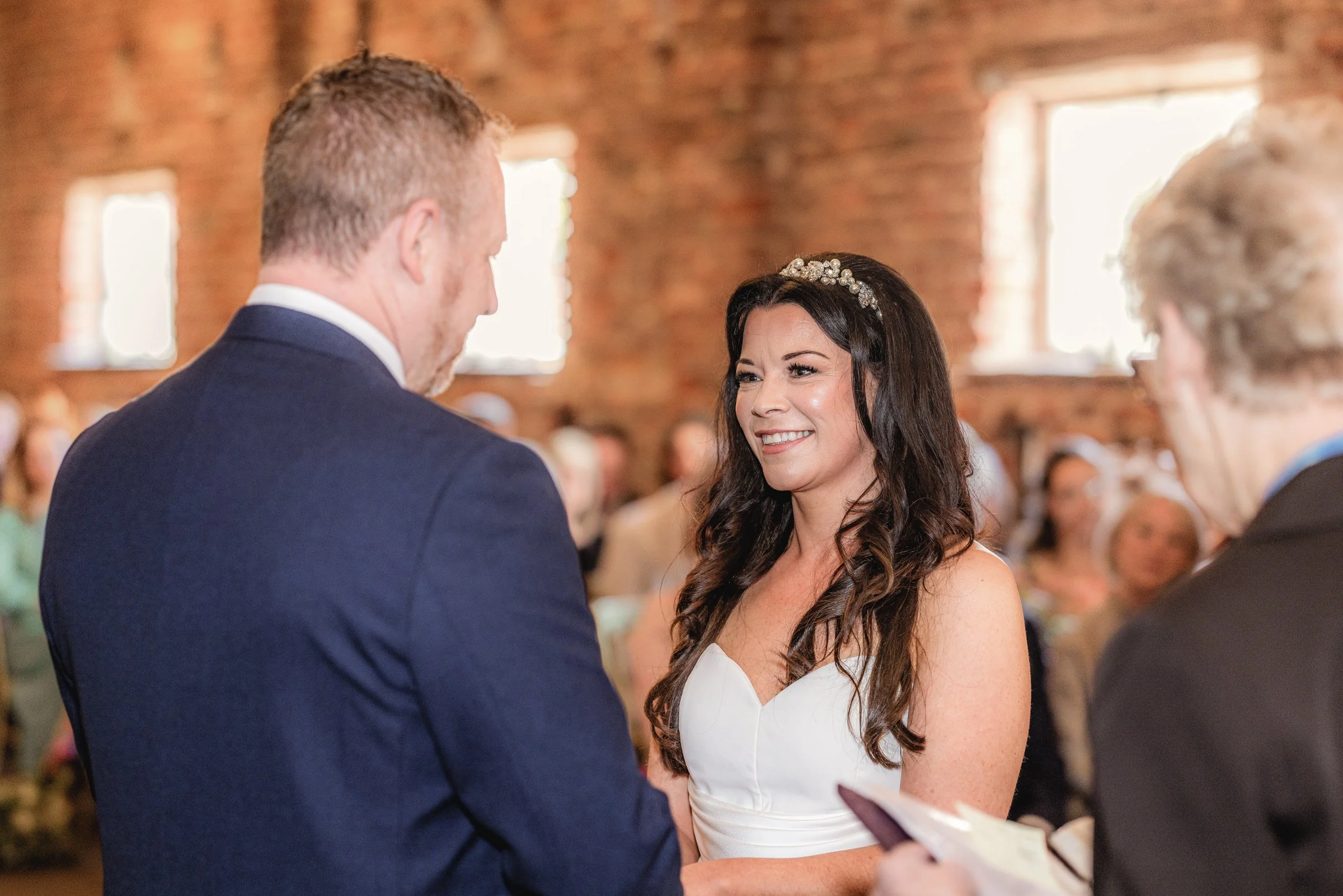 A bride and groom exchanging vows during a wedding ceremony inside a rustic venue with exposed brick walls and windows.