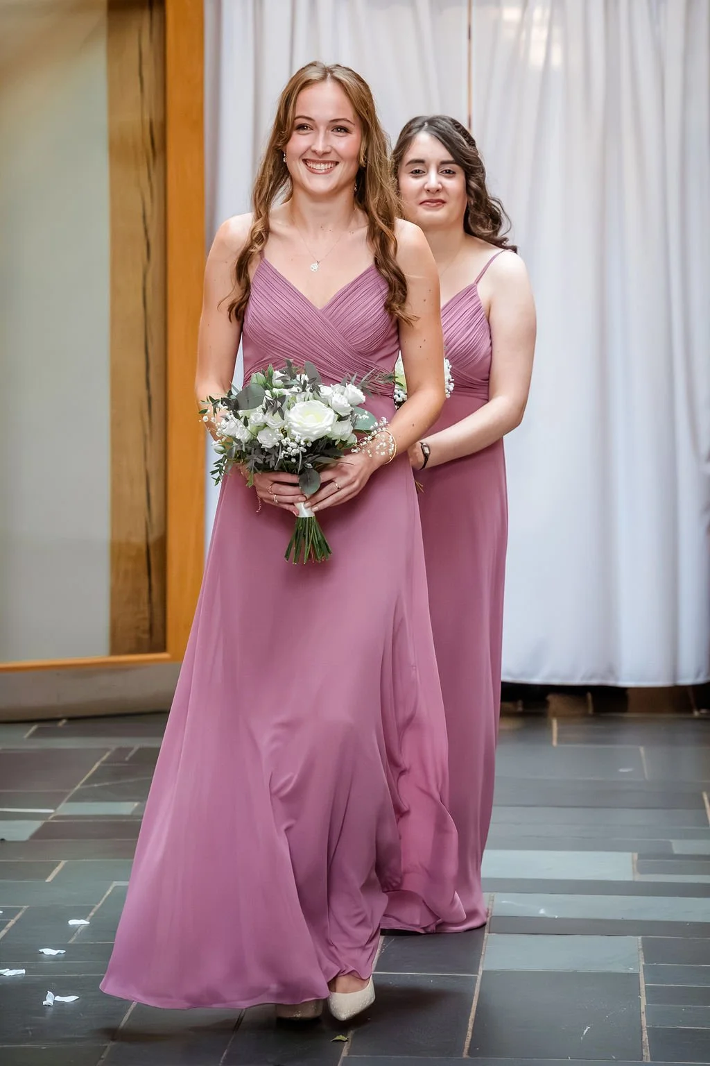 Two women in matching mauve bridesmaid dresses, one holding a bouquet of white roses and greenery, standing indoors with curtains and a wooden beam background.
