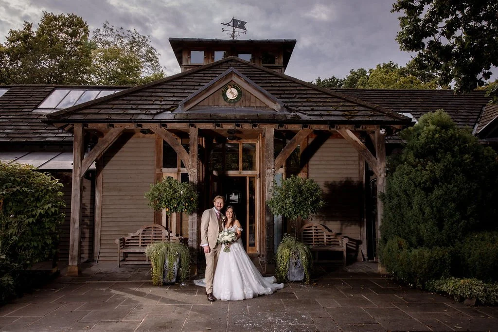 A newlywed couple stands in front of a rustic building with wooden beams, two benches, and lush greenery. The bride wears a white wedding gown, and the groom is in a light-colored suit.