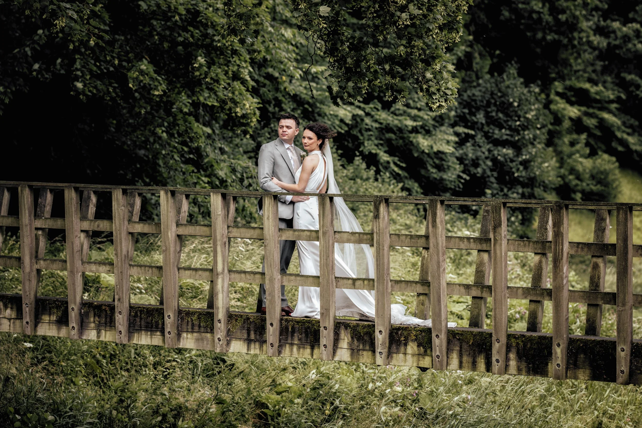 A bride and groom standing on a wooden bridge surrounded by green trees.