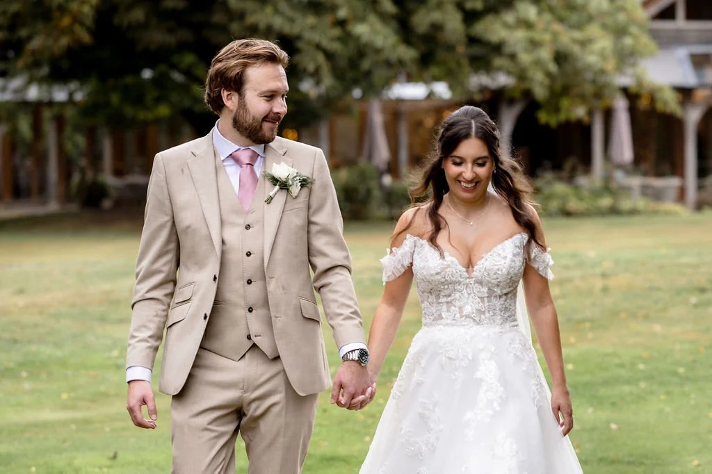 A bride and groom walking hand in hand outdoors, smiling, with the bride wearing a white wedding dress and the groom in a beige suit with a pink tie, surrounded by greenery.