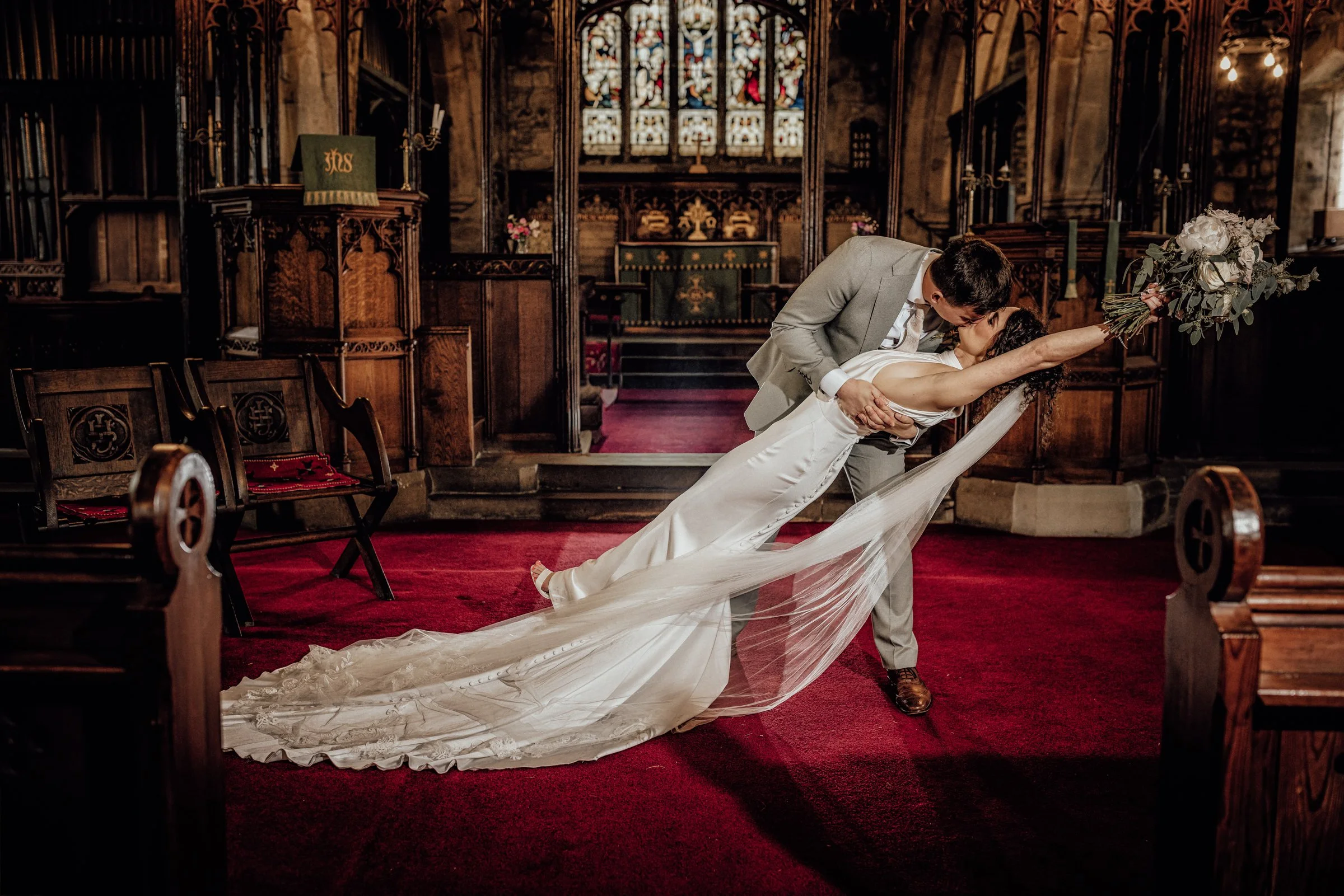 A bride and groom kissing inside a church, with the groom dipping the bride. The bride holds a bouquet of white flowers in one hand and the other around the groom's neck. The church has wooden pews and stained glass windows.