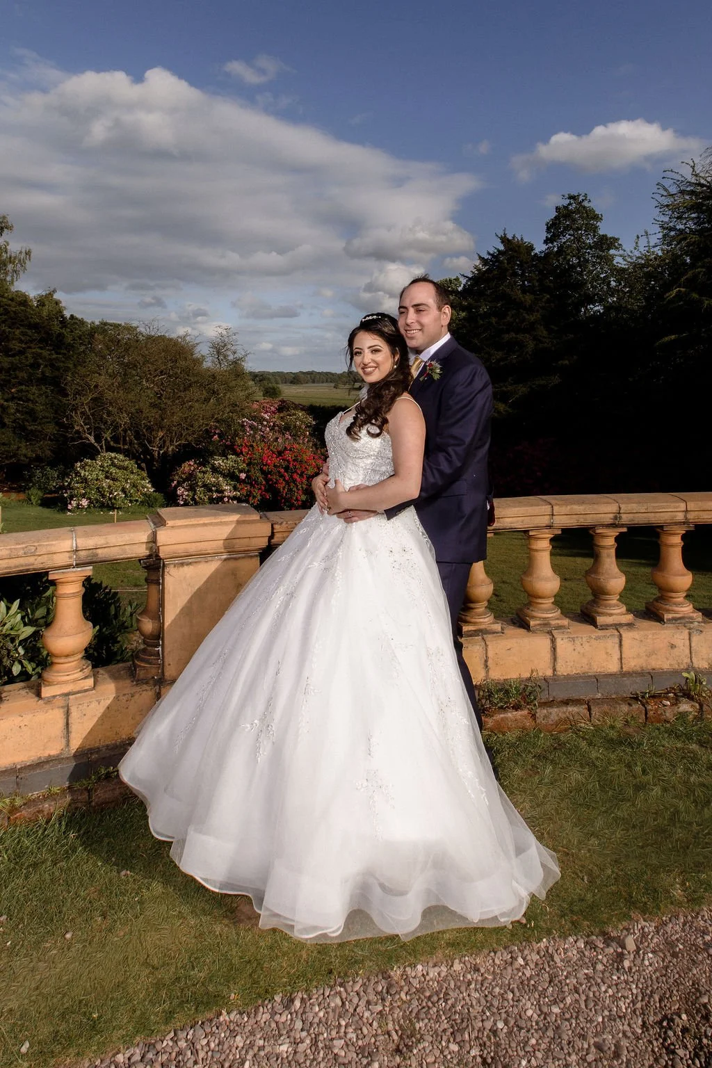 A bride and groom standing outside on a stone balcony, with trees and a blue sky with clouds in the background, smiling for a wedding photo.