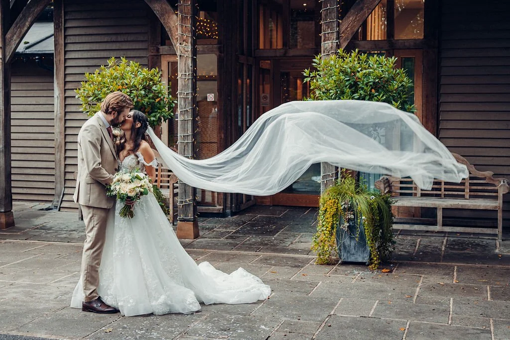 Wedding couple kissing outdoors, with bride in a white gown holding a bouquet, groom in a beige suit, and a flowing veil in the air.