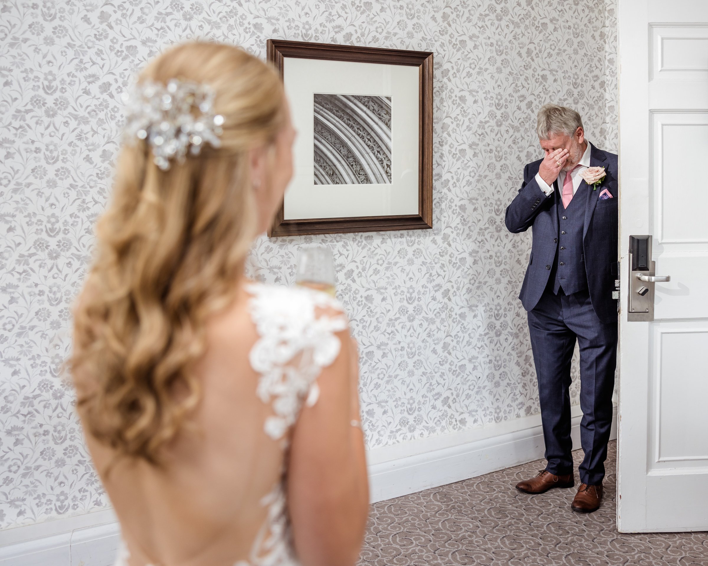 A bride with long, curly blonde hair and a floral hairpiece looks at a man in a suit who is crying and covering his face, standing in a doorway.