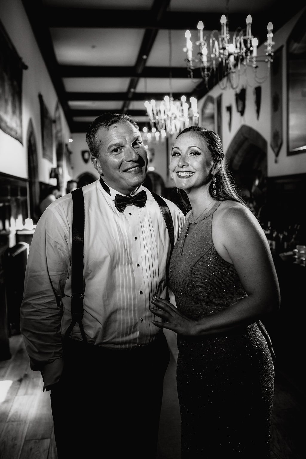 A black and white photo of a man in a tuxedo with suspenders and a woman in a sparkly dress, both smiling, at a formal event in an elegant room with chandeliers.