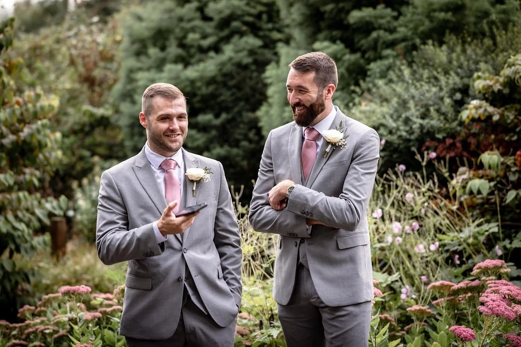 Two men in gray suits with pink ties and white boutonnieres standing outdoors among pink and purple flowers with greenery in the background, smiling and talking.