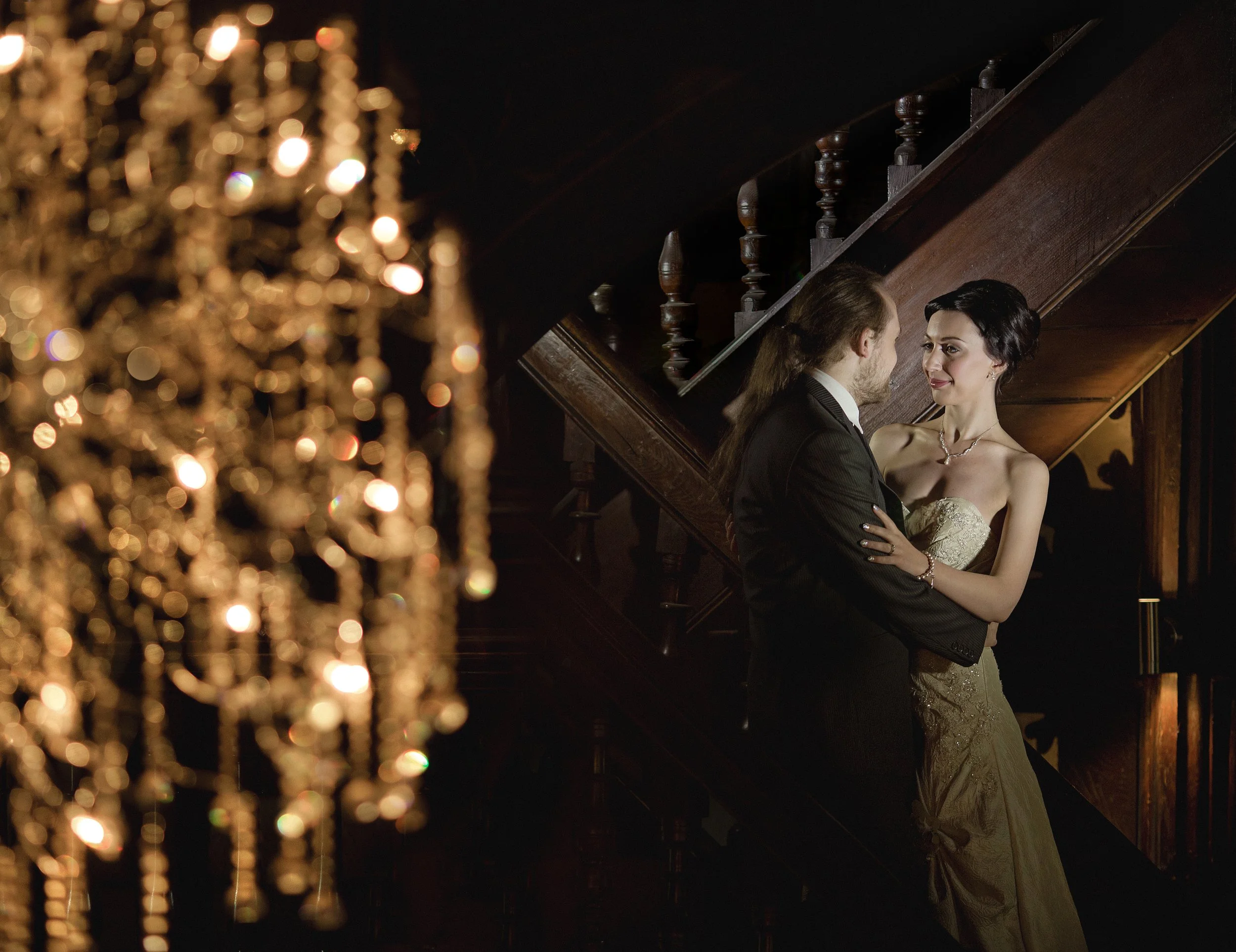A man and woman in formal attire standing on a staircase, gazing at each other in a dimly lit, elegant setting with a chandelier partially visible on the left.