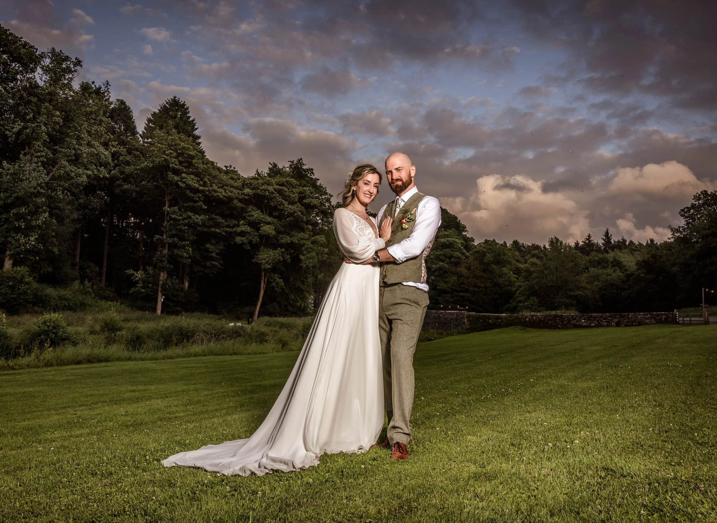 A bride and groom stand together on a grassy field during dusk, smiling with trees in the background and a partly cloudy sky overhead.