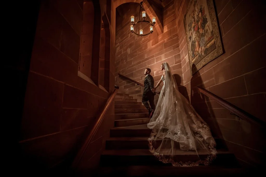 A bride and groom holding hands on a wooden staircase inside a dimly lit, stone-walled building, with a tapestry hanging on the wall, a chandelier overhead, and the bride's long lace train flowing behind her.