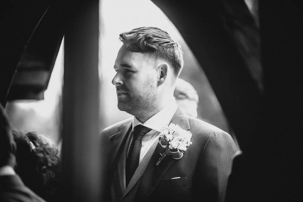 A man in a suit with a boutonniere, looking to the side, taken in black and white.