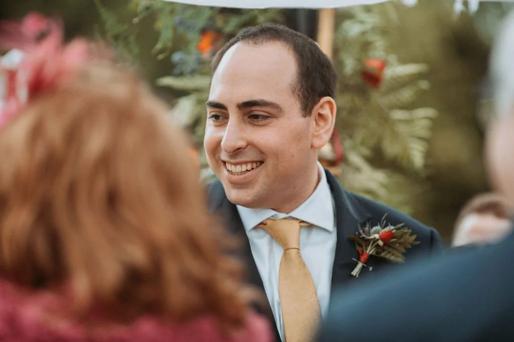 A man with short dark hair and a light complexion, dressed in a suit with a boutonnière, smiling at a conversation during a wedding or formal gathering.