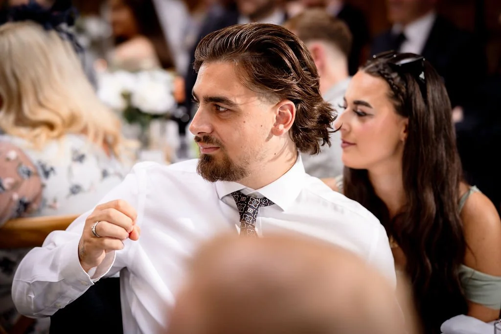 A young man with long brown hair and a beard wearing a white shirt and a patterned tie, seated at a formal event, with a woman with dark hair and sunglasses resting on her head sitting behind him.