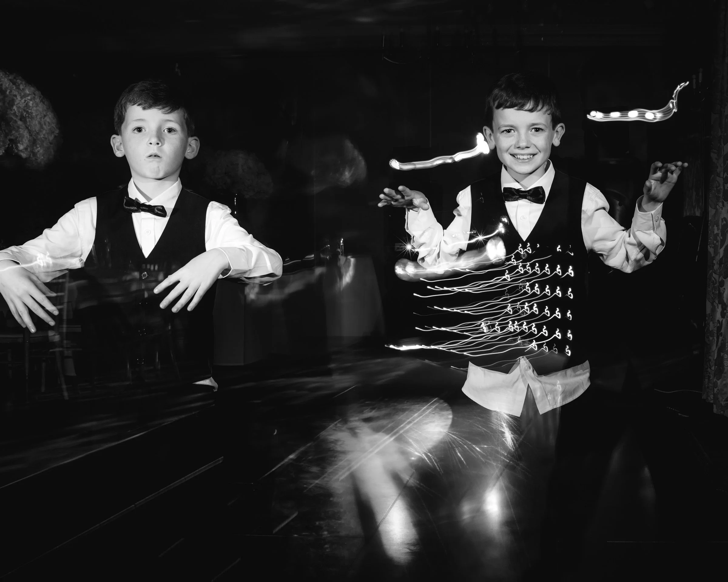 Two boys in tuxedo vests and bow ties standing in front of a mirror, one with a serious expression and the other smiling, with light trails around them creating a dynamic effect.