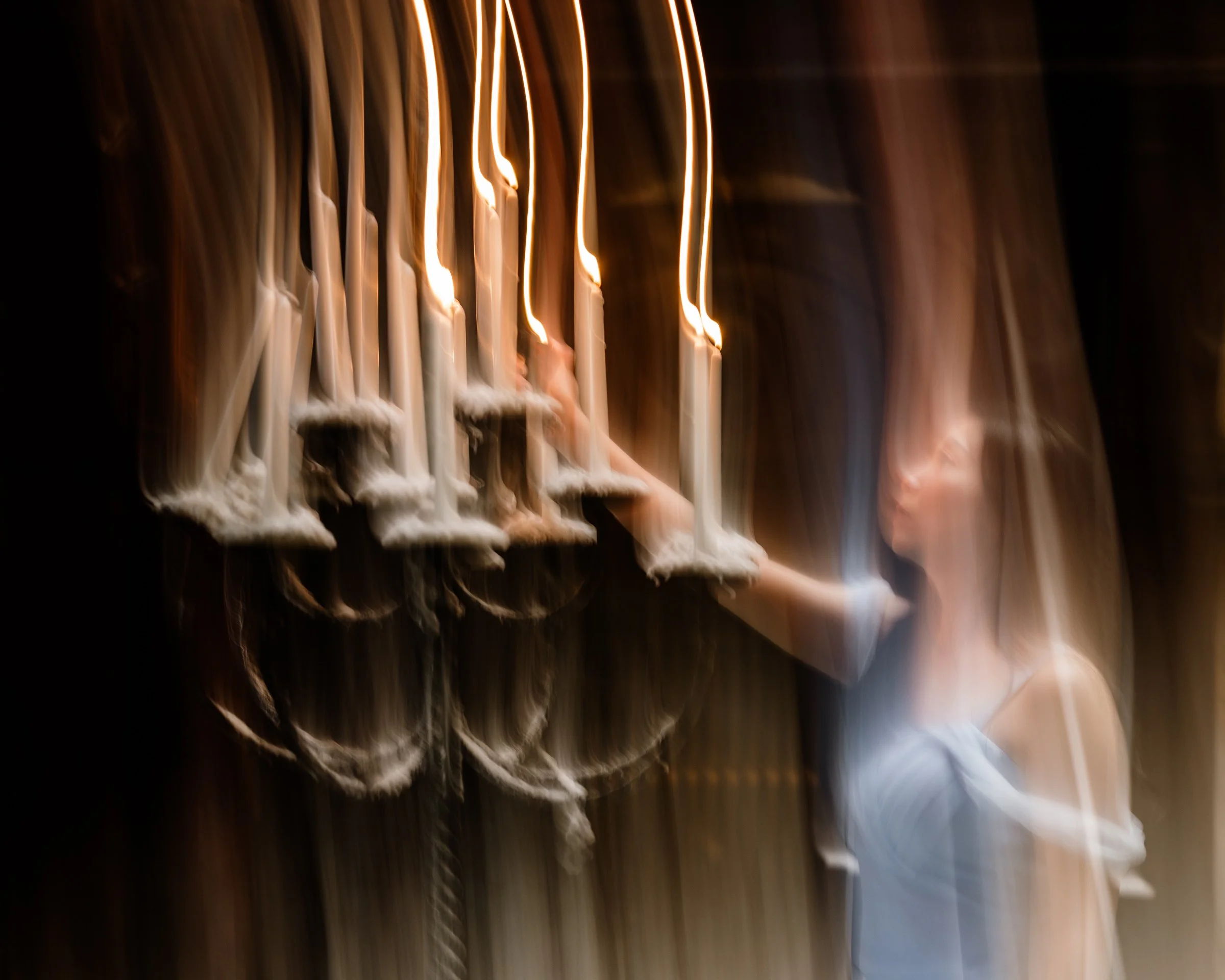Blurred picture of candles with flames, with a dark background and some figures or objects in the background.