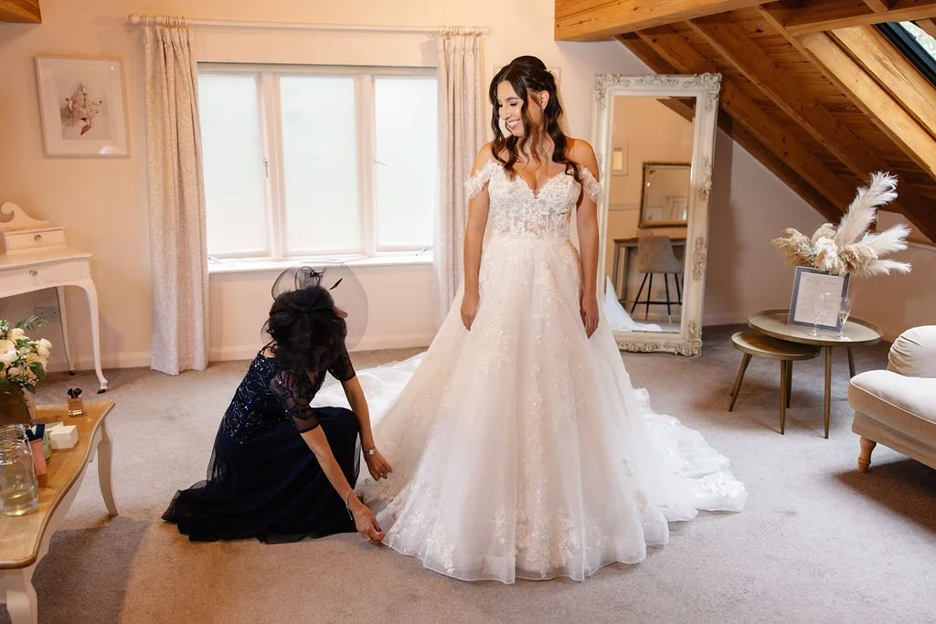 A woman in a wedding dress standing in a bright room while another woman adjusts the dress.
