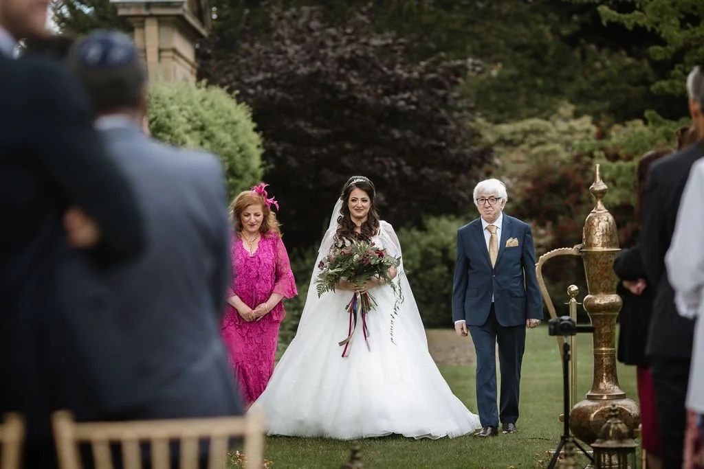 Bride in white wedding gown holding a bouquet, walking with two adults, outdoors in a garden setting, surrounded by guests.