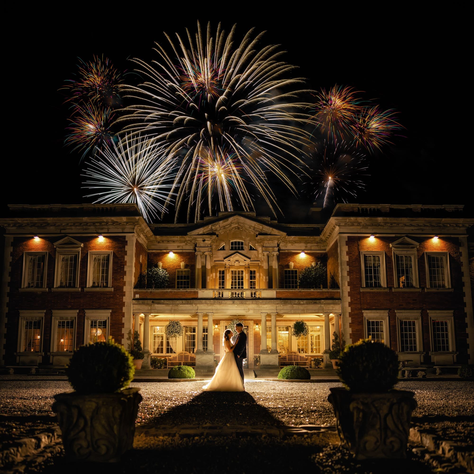 A bride and groom holding hands and dancing in front of a large house during fireworks display at night.