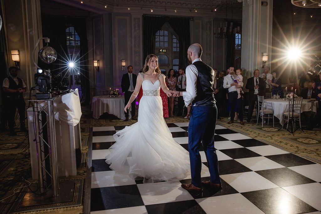 A bride and groom dancing on a checkered dance floor at their wedding reception with guests watching in an elegant venue with high ceilings and large windows.