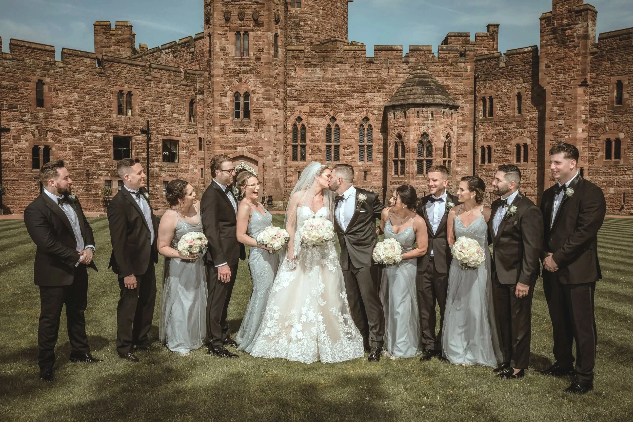 A wedding party standing on a lawn in front of a large castle, with the bride and groom kissing in the center. The group includes bridesmaids, groomsmen, and the bride in a white gown with lace details and a veil, and the groom in a black tuxedo. Bridesmaids wear light gray dresses, and groomsmen wear black tuxedos with bow ties, all holding bouquets of flowers.
