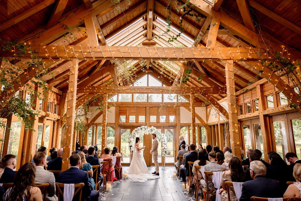 Wedding ceremony inside a wooden lodge with large windows, where a bride and groom stand under an archway decorated with white flowers.