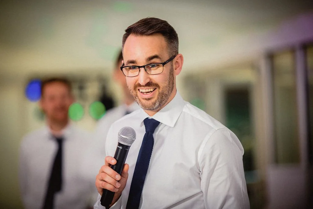 A man with glasses and a beard is smiling and holding a microphone, standing in an indoor setting with blurred people and colorful lights in the background.