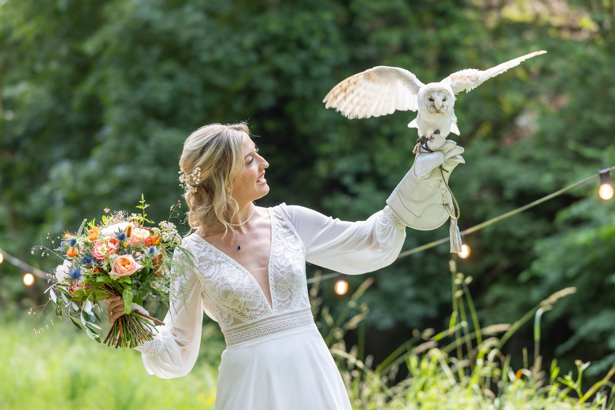 A woman in a white lace dress holding a bouquet of flowers in one hand and a white owl on her gloved hand in the other, outdoors with greenery and string lights.