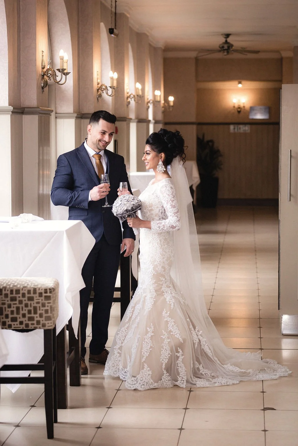 A bride and groom smile at each other while holding glasses and a wedding bouquet in a warmly lit reception hall.