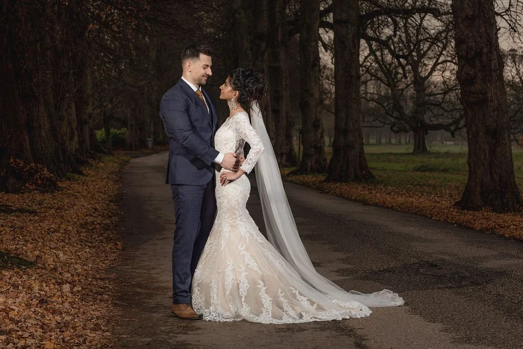 A bride and groom standing close together on a tree-lined road during sunset, with the bride in a lace wedding gown and veil and the groom in a dark suit, appearing happy and romantic.