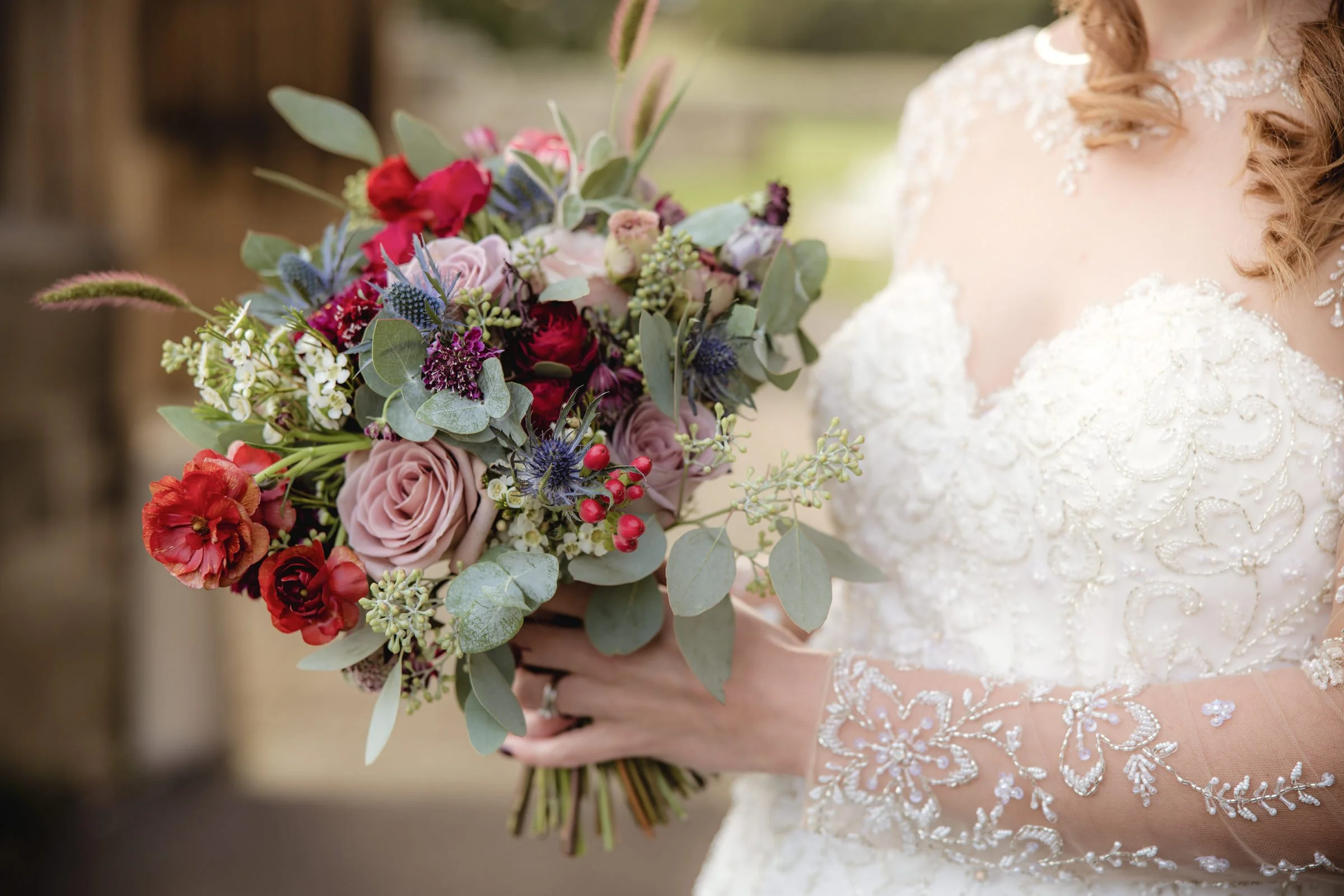 A bride holding a colorful bouquet of flowers, including roses, berries, and greenery, while wearing a lace wedding dress with detailed embroidery.