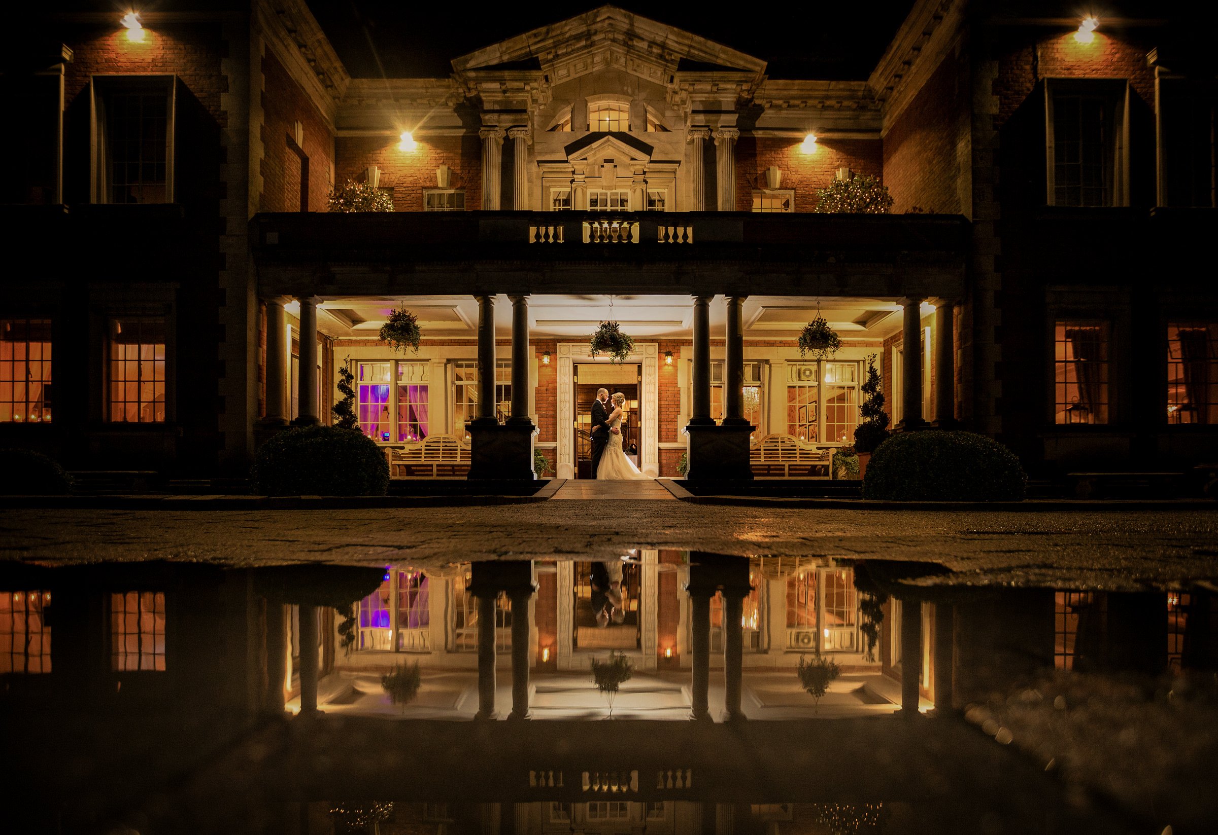 A couple in wedding attire sharing a kiss at dusk in front of a large, illuminated mansion with a reflection in a puddle.