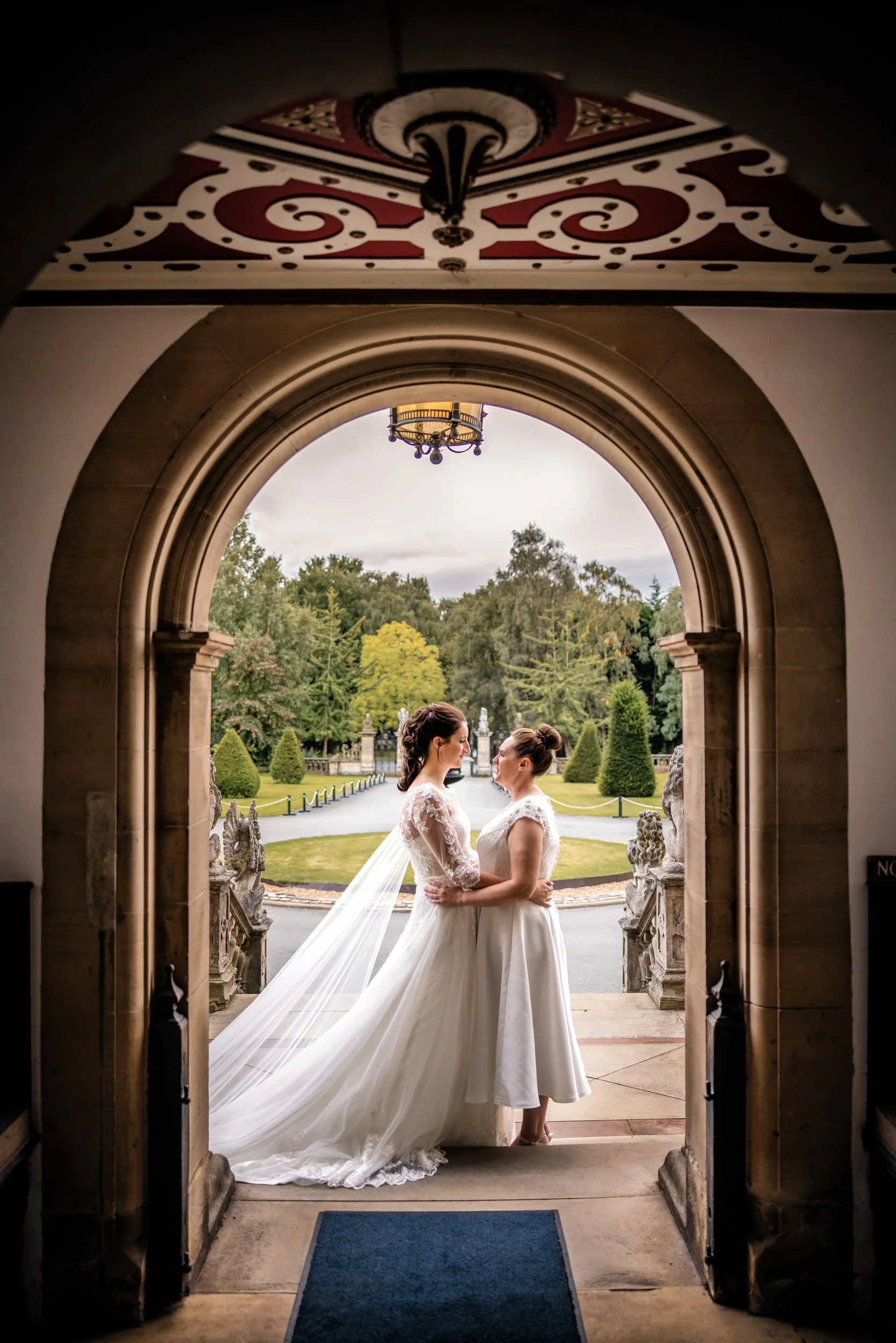 Two women in wedding dresses standing face to face at the threshold of a stone archway, holding each other, with a garden and trees in the background.