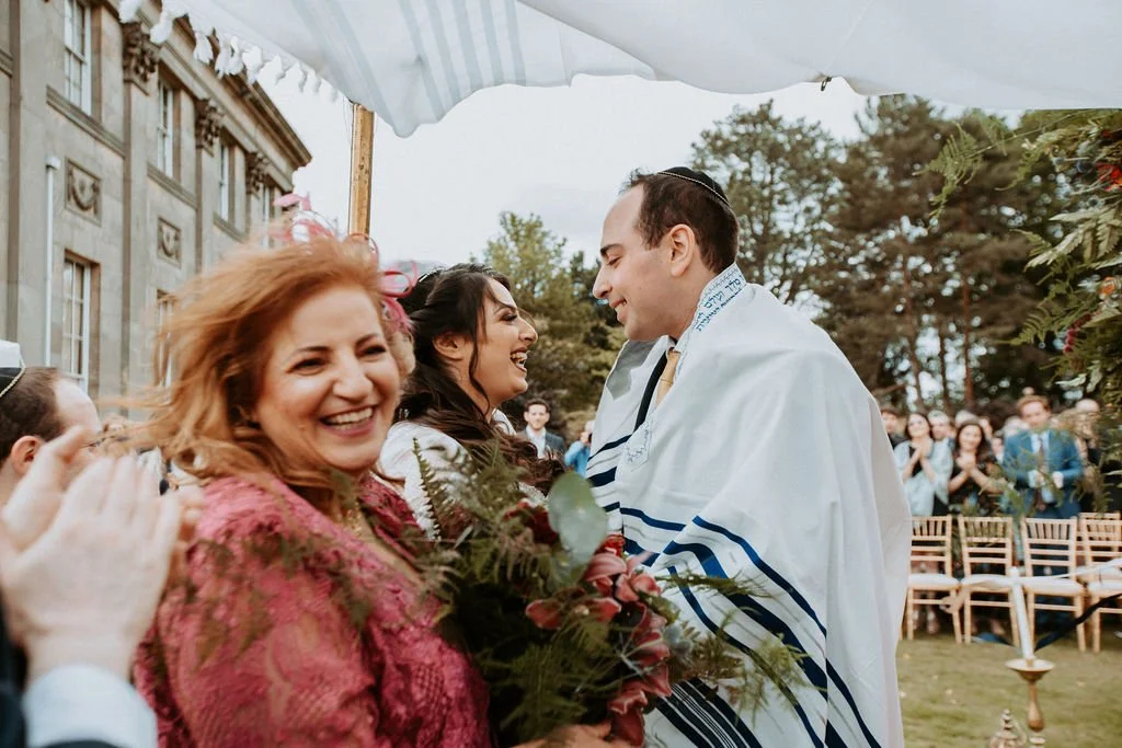 A wedding ceremony outdoors with a smiling bride and groom facing each other, surrounded by friends and family, some in the background clapping. The bride holds a bouquet of flowers, and there's a historic building in the background.