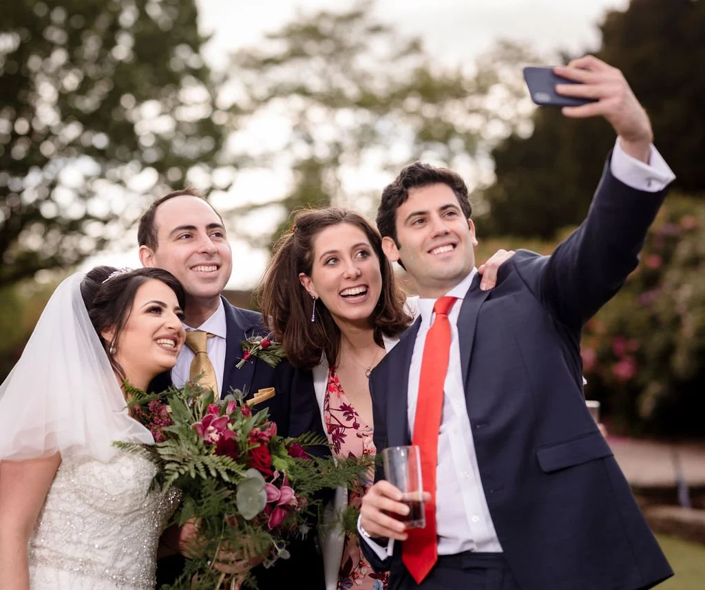 A group of four people, including a bride in a wedding dress holding a bouquet and three others in formal clothing, taking a selfie outdoors during a wedding celebration.