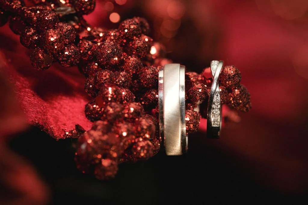 Silver wedding bands and a diamond ring resting on red glittery berries and red fabric.