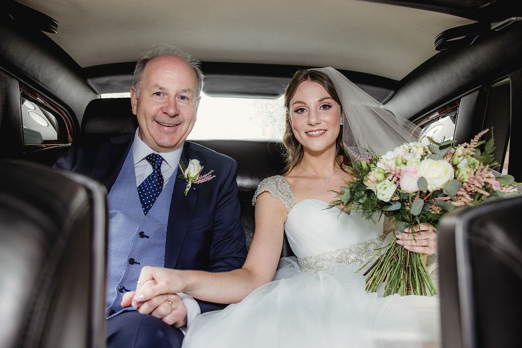 A bride and an older man, likely her father, sitting inside a car. The bride is holding a bouquet of flowers and wearing a white wedding dress with a veil. They are smiling and holding hands.