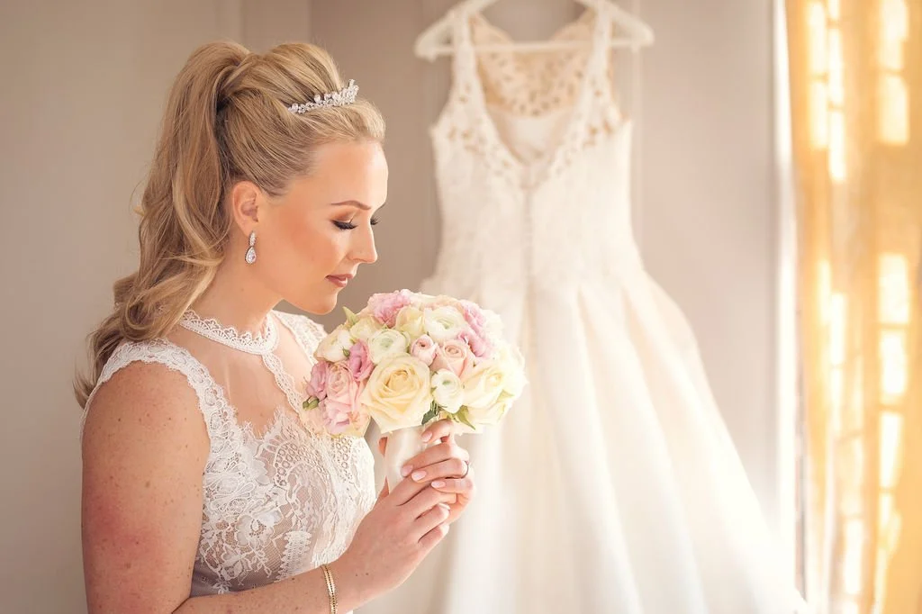 Bridal bride holding a bouquet of pink and white roses, standing near her wedding dress hanging in the background during daylight.