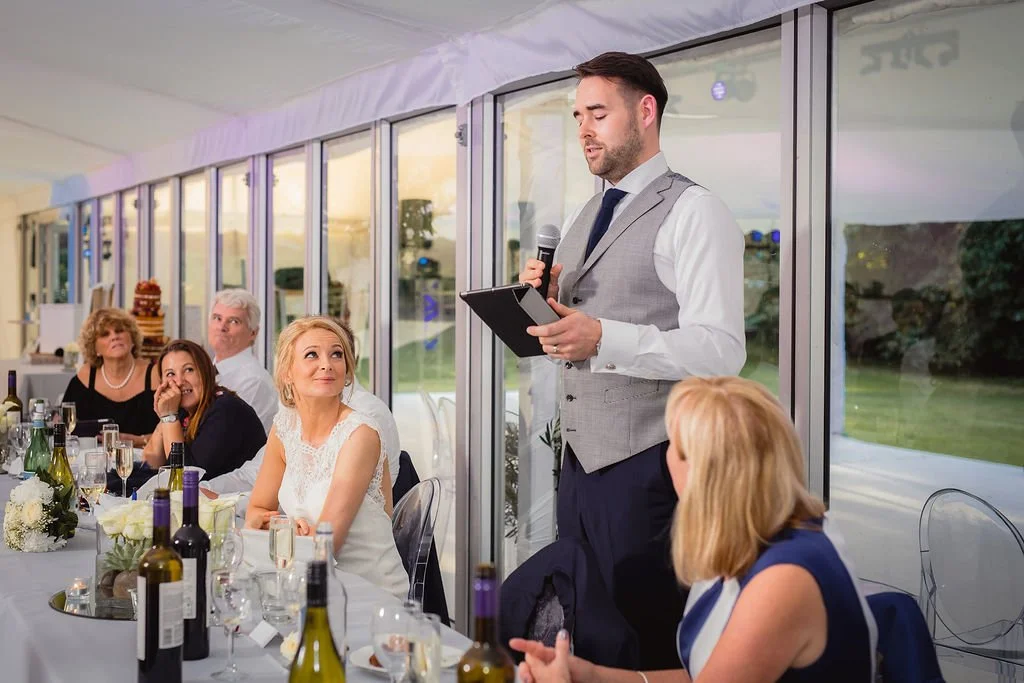A man giving a speech at a wedding reception, standing next to a table with seated guests, some of whom are looking at him and smiling.
