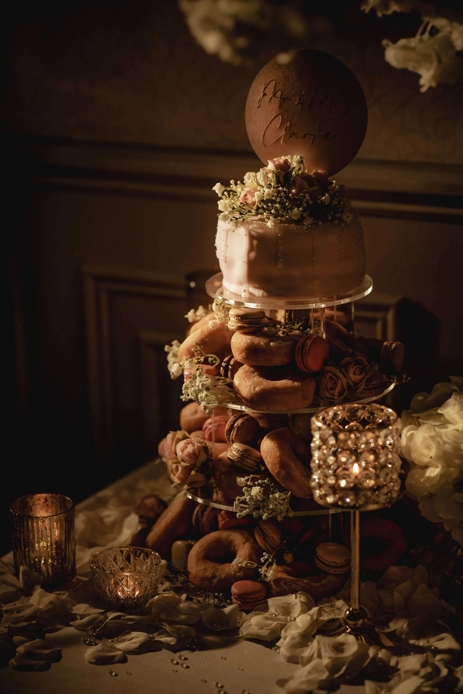 A three-tier wedding cake decorated with flowers and a wooden topper with handwritten names, surrounded by macarons, donuts, roses, and candles on a table scattered with rose petals, in a dimly lit setting.
