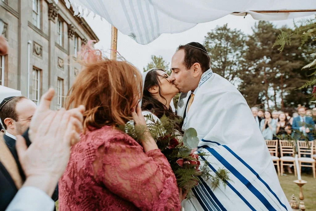 A couple is kissing at a wedding ceremony, with the groom wrapped in a prayer shawl, and guests clapping in the background.