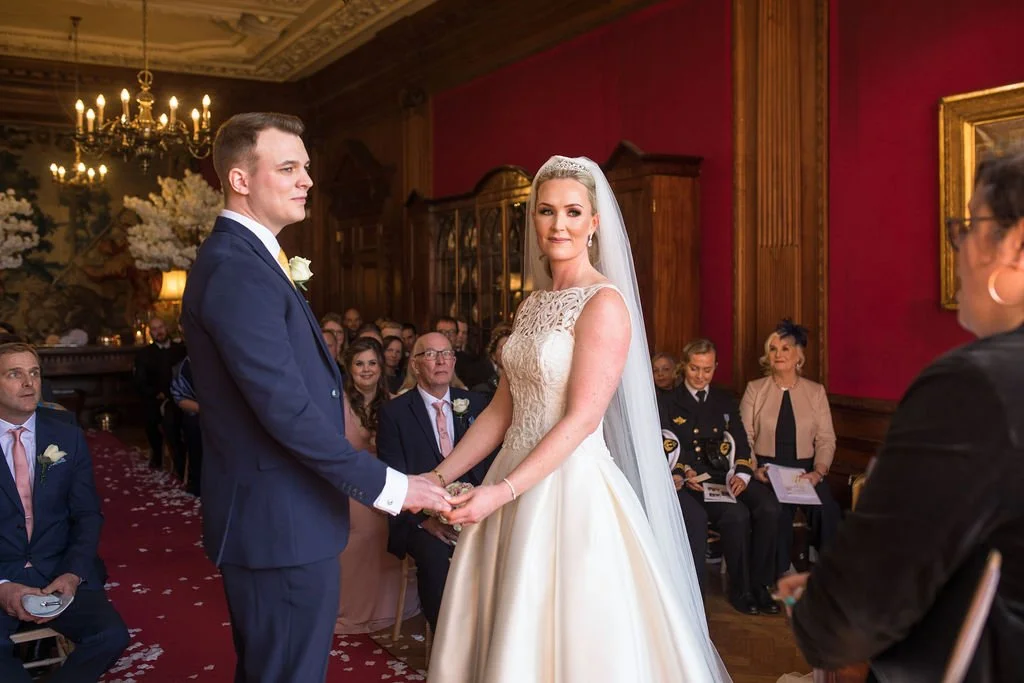 A bride and groom holding hands during their wedding ceremony in an elegant, historic room with dark wood paneling, red walls, chandeliers, and seated guests watching.