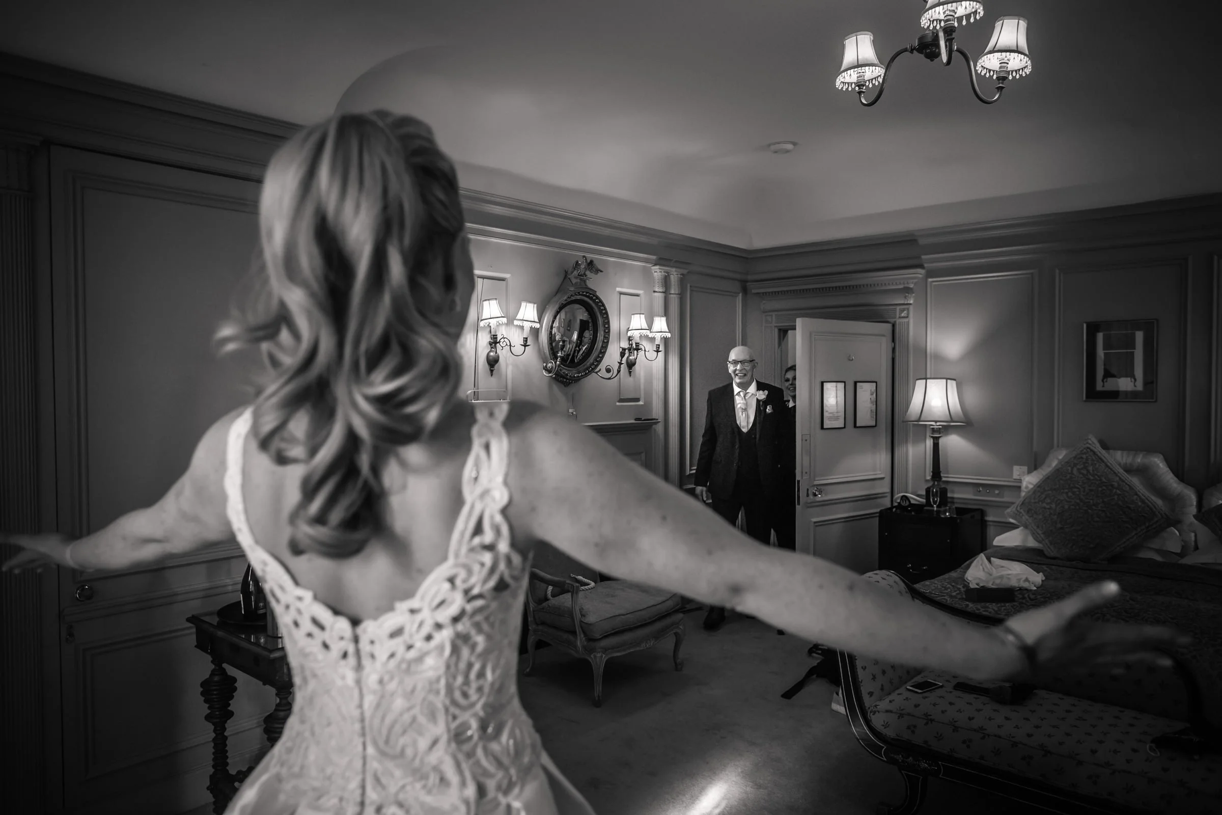 Black and white photograph of a bride with arms outstretched, facing an older man in a suit in a warmly decorated room.
